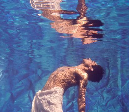 A shirtless man swimming underwater in a pool wearing a cloth skirt has his face looking up towards the sky,