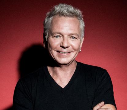 A white man in his 60s with grey hair wears a black t-shirt and has his arms crossed, standing in front of a red background.