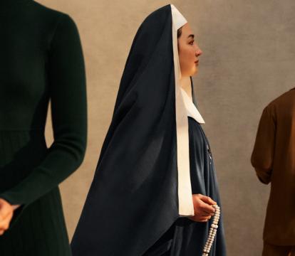 A woman dressed as a nun faces the right. Another woman wearing black holds a candle and a man faces the back wearing a copper coloured suit.