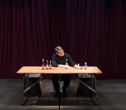 A person sits alone at a wooden table in a dimly lit room with deep red curtains. Bottles are organized on shelves against the walls, creating an intimate, contemplative mood.
