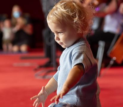 A toddler in a blue sweater and floral skirt dances on a red carpeted floor with people and musicians playing string instruments in the background.