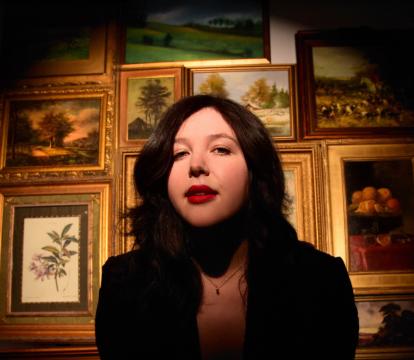 A woman with brown hair looks at the camera standing in front of a wall of gold picture frames.