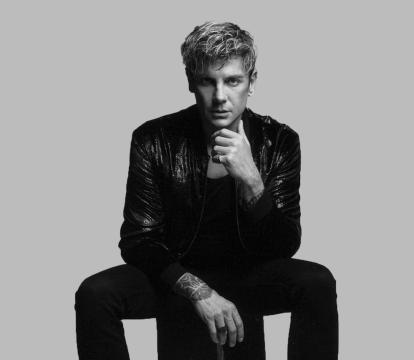 A black and white image of a white man with blonde, spikey hair sitting on a stool wearing a leather jacket