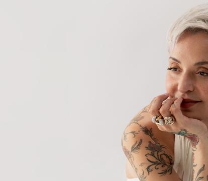 A woman adorned with tattoos with short, bleached blonde hair is comfortably positioned on a chair, resting her chin in her hand.
