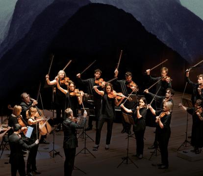 A group of musicians mid-performance on a stage collaged into the side of a mountain with dark mist and clouds in the background.