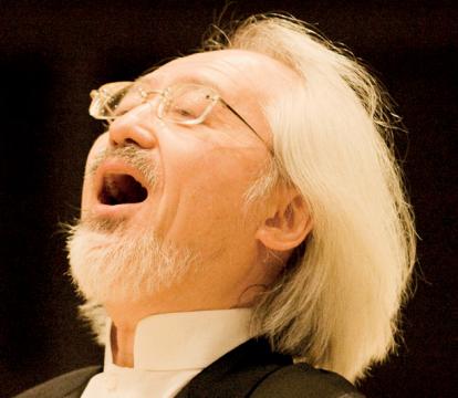 A man with white hairs and in black suit singing on the stage.