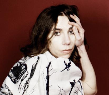 A white woman with long brown hair wears a stylish white dress with a black splash pattern. She's crouched down and looking at the camera.
