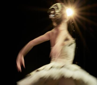 A ballerina wears a gold tutu and hairpiece. Her back is to the camera and her arms are stretched behind her, mid-dance move. A spotlight is seen behind her.