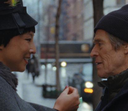 A woman wearing a coat and a hat with a feather in it stands on an urban street facing a man who is wearing a beanie and coat.