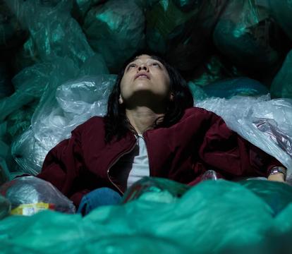 A person in a grey shirt and red jacket is looking upward while seated on and surrounded by full garbage bags.