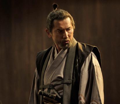 A man in a traditional Japanese garment is standing while other men dressed similarly are  crouched and surrounding him. A helmet and armour is visible in the background.