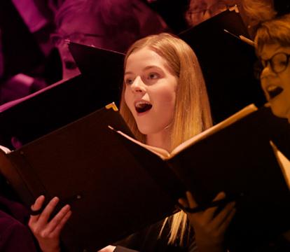 A blonde girl with an open song book sings amongst a choir