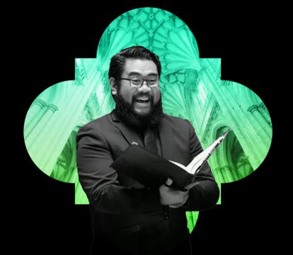 Man with a beard in a black suit sings with a cathedral as the background