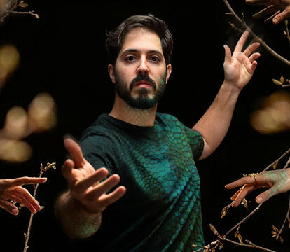 A man stands with one arm reaching forward and one back. He is surrounded by tree branches and other hands.