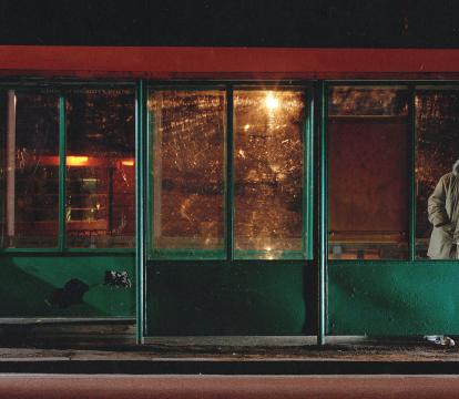 A man in a thick coat leans against an empty bus stop at night.