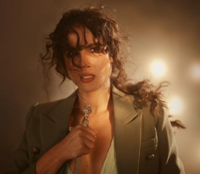 A white woman with a messy, curly ponytail and wearing a beige blazer looks worried at the camera while holding the handle of a sword.