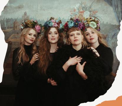 Four women in dark dresses stand closely, each wearing a floral crown.