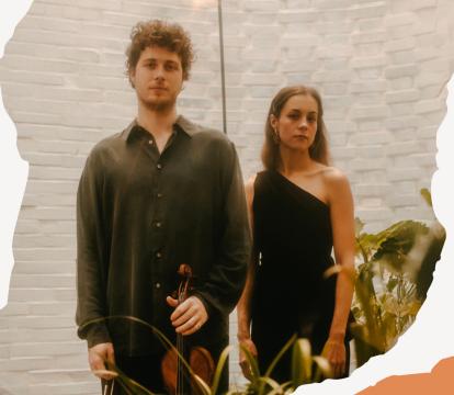 A man and woman stand side by side in a bright room, surrounded by plants. The man holds a violin.