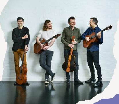 Four men holding guitars stand casually against a white brick wall. They are smiling and interacting.