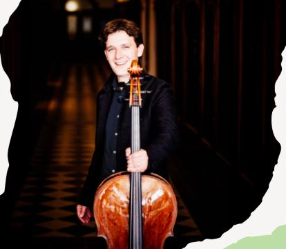 A white man smiles while holding a cello in a dimly lit hallway.