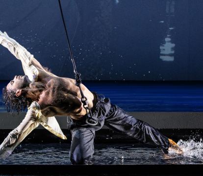 A white man with grey hair in a suit stands looking at a male dancer knelt on the floor. The dancer wears a harness and is dancing in water.