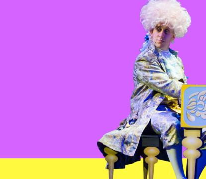 A man wearing a Mozart costume and a big white wig sitting at a grand piano