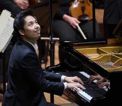 A Korean man wearing a black suit sits at a grand piano looking up and smiling