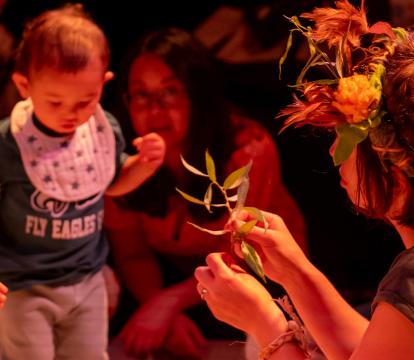 A women wearing a crown of flowers is holding up some leaves to two toddlers.