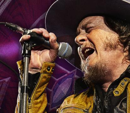 A man in yellow jacket and a cow boy hat singing in to the mic.