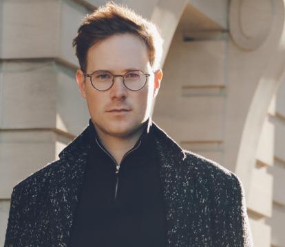 A portrait of a man with glasses in a dark shirt and coat standing in front of a stone building on a sunny day.