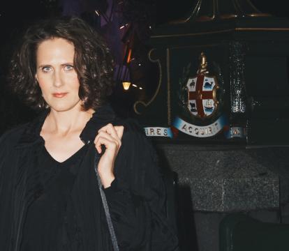 A women standing on a dark street at night, wearing black, with an instrument case over her shoulder.