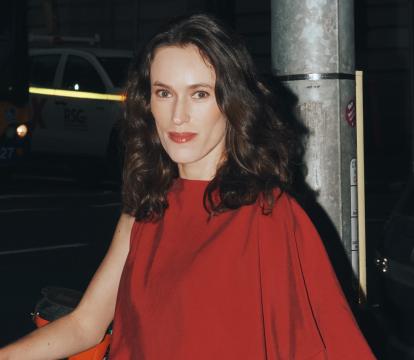 A woman wearing a red asymmetrical dress outside at night with the back of a dark grey car in the background.