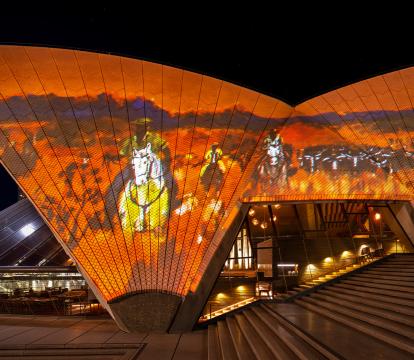 The Sydney Opera House sails at night, illuminated with an Australian outback landscape in orange and browns.