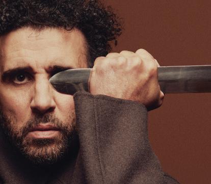 A man with stubble beard and curly hairs holding a sword blade.