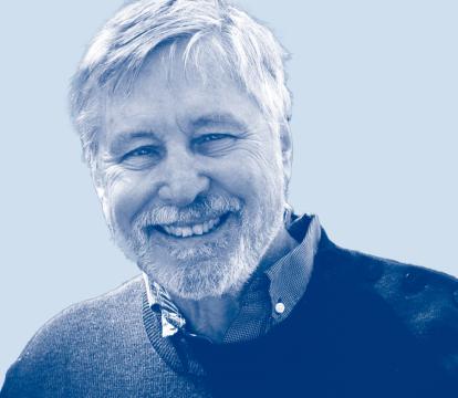 A white man with white hair and a beard smiling. He wears a blue sweater.