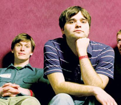 A group of four men seated on red chairs in front of a pink background.