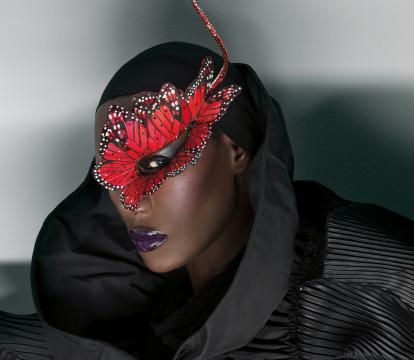 A black woman wearing a striking red butterfly mask adorned with white dots, set against a dark, textured outfit with a high collar.