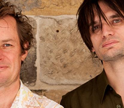 Two men in short-sleeved shirts standing outside a brick wall.