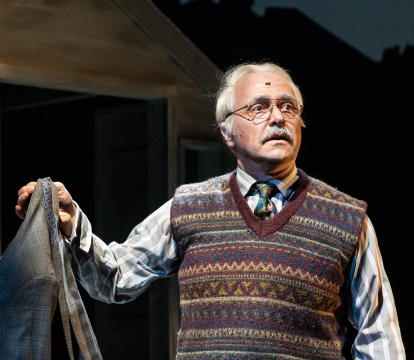 An older man with grey hair and a moustache stands wearing a wool vest, shirt and tier holding a blazer jacket. He is on a set that resembles a backyard at night.