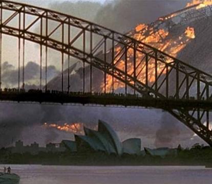 The Harbor bridge and Sydney Opera House in front of a large object on fire.
