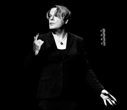 A person in a dark suit gestures passionately onstage in a spotlight against a black background, conveying intensity and focus.