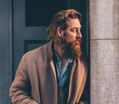 A man with brown hair and a long brown beard wearing a camel-colored coat over a denim shirt, leans on a door frame.