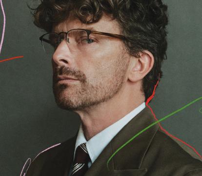 A man wearing glasses and a dark green suit looking sidelong against a grey-green background with subtle scribbled marks in pink, red and green.
