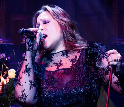 A Filipino-Australian woman with brown layered, shoulder-length hair holds a microphone in her right hand and the microphone stand in her left hand. She's singing on stage in front of an orchestra.