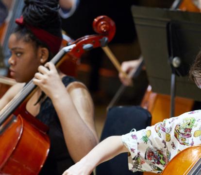 Two rows of young people seated in a room, playing cellos.
