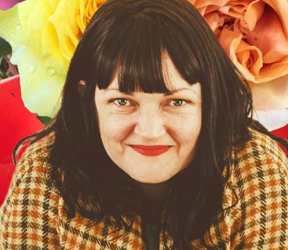 A lady with dark hair, a fringe and red lipstick infront of a floral background