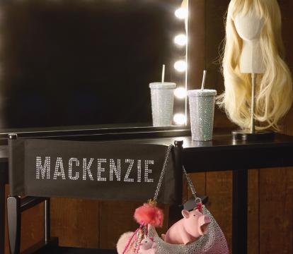A long blonde wig and cup with a straw sit on a black dressing table with a mirror edged with lights and a folding chair with Mackenzie on the back and a pink handbag hanging off one side.