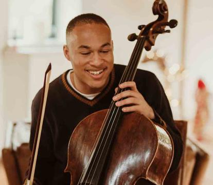 Sheku Kanneh-Mason holds a Cello in front of a piano.