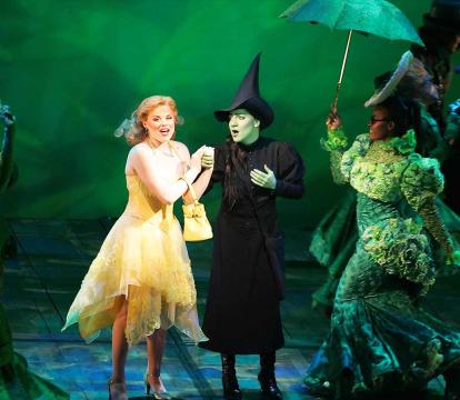 Actors on stage including a witch with a green face, a blonde woman in a yellow dress and a woman in a green dress.