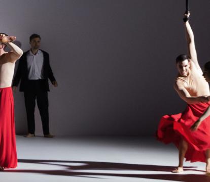 Two cast members wearing long red skirts, one is horizontal in the air attached to an aerial rope, the other assisting her movement
